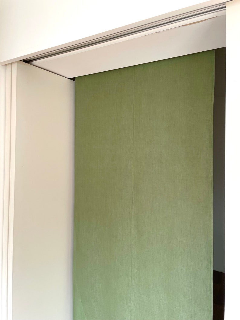 Puede incluir: Un divisor de habitaci&oacute;n de tela verde cuelga de un sistema de rieles. El divisor est&aacute; hecho de una tela texturizada y est&aacute; parcialmente abierto, revelando una pared blanca detr&aacute;s.