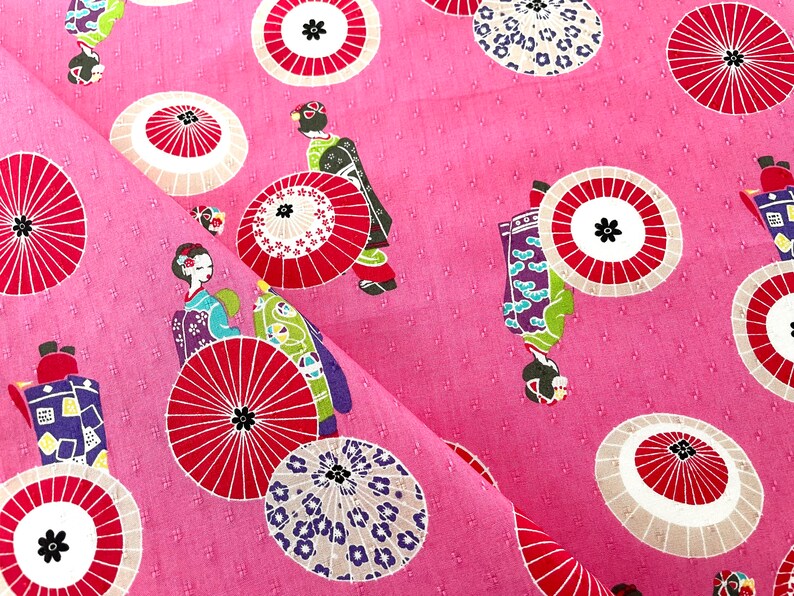 May include: Pink fabric with a repeating pattern of geishas and red and white umbrellas. The umbrellas have a white background with red spokes and a black center.