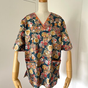 Puede incluir: Una bata de trabajo azul verdoso con cuello en V y mangas cortas. La tela presenta un estampado floral colorido con detalles dorados. El cuello y los bolsillos tienen un ribete naranja contrastante.