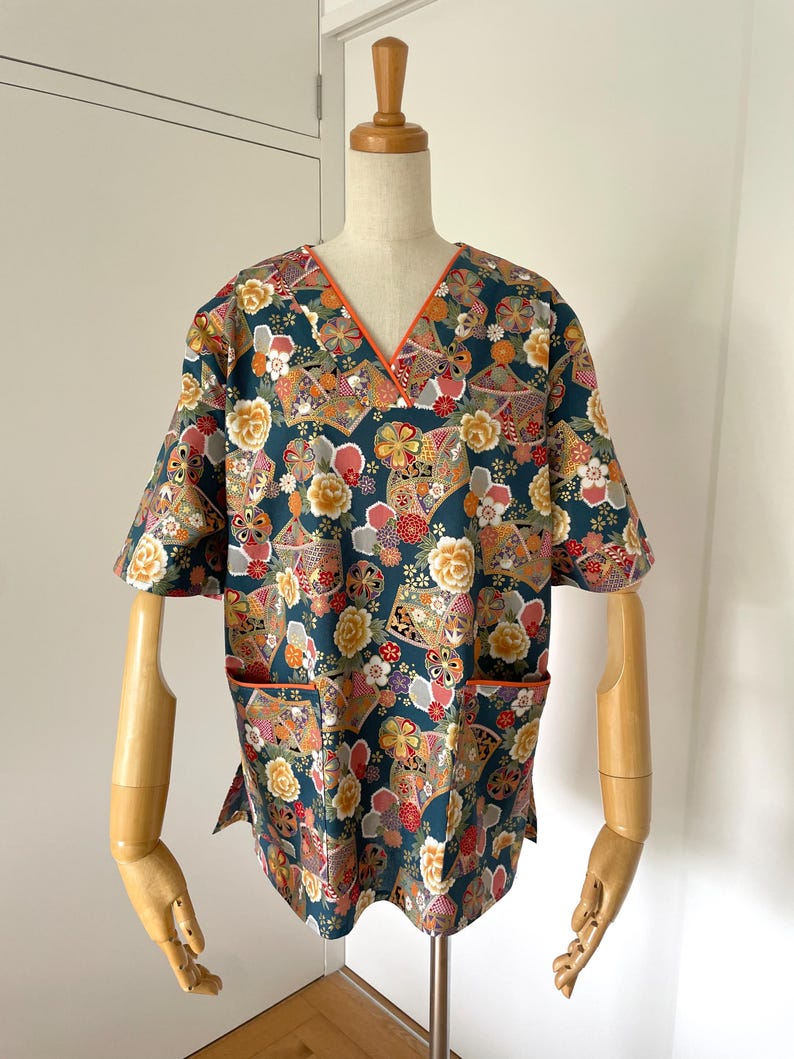 Puede incluir: Una bata de trabajo azul verdoso con cuello en V y mangas cortas. La tela presenta un estampado floral colorido con detalles dorados. El cuello y los bolsillos tienen un ribete naranja contrastante.