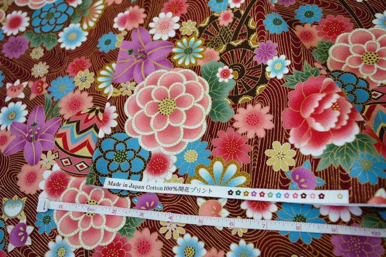 May include: A dark red cotton fabric featuring a floral pattern with pink, blue, and purple flowers, and gold line details. A white measuring tape shows a length of 10 inches. The fabric edge includes the text Made in Japan Cotton 100% and flower icons.