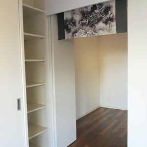 Puede incluir: Vista interior de una habitaci&oacute;n con una estanter&iacute;a blanca empotrada a la izquierda. Una puerta corredera revela una habitaci&oacute;n con suelo de madera oscura y un rollo decorativo con un dise&ntilde;o de drag&oacute;n en blanco y negro.