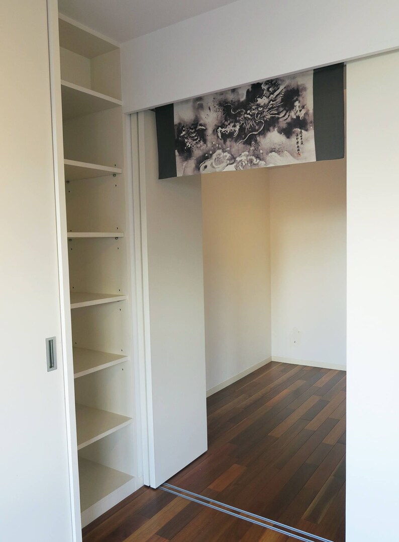 Puede incluir: Vista interior de una habitaci&oacute;n con una estanter&iacute;a blanca empotrada a la izquierda. Una puerta corredera revela una habitaci&oacute;n con suelo de madera oscura y un rollo decorativo con un dise&ntilde;o de drag&oacute;n en blanco y negro.