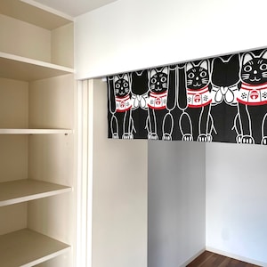Puede incluir: Cortina japonesa Maneki Neko en blanco y negro. La cortina presenta m&uacute;ltiples figuras de gatos con patas levantadas, collares rojos y detalles en blanco. La cortina cuelga en una habitaci&oacute;n blanca con una estanter&iacute;a empotrada.