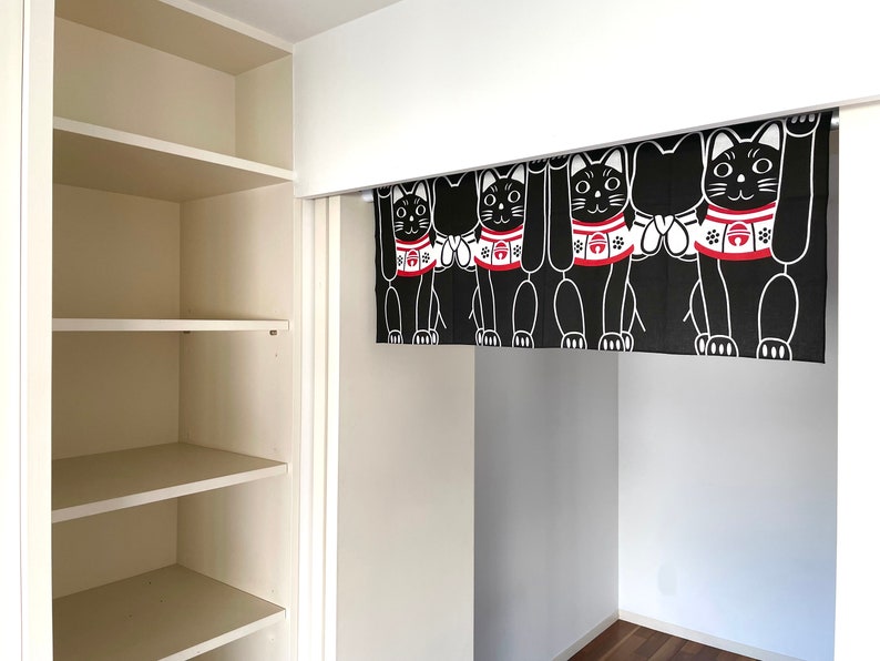 Puede incluir: Cortina japonesa Maneki Neko en blanco y negro. La cortina presenta m&uacute;ltiples figuras de gatos con patas levantadas, collares rojos y detalles en blanco. La cortina cuelga en una habitaci&oacute;n blanca con una estanter&iacute;a empotrada.