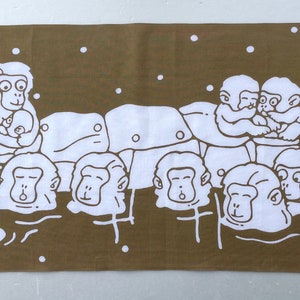 May include: A brown rectangular towel featuring a white illustration of several monkeys. The monkeys are depicted in a hot spring, with some holding babies. White dots are scattered across the background.
