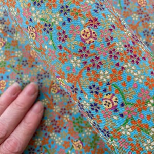 Puede incluir: Una tela azul claro con un patr&oacute;n denso y repetitivo de peque&ntilde;as flores de cerezo naranjas, blancas y moradas, tallos verdes y motivos decorativos circulares en tono dorado con detalles geom&eacute;tricos complejos en todo el tejido.