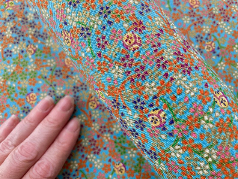 Puede incluir: Una tela azul claro con un patr&oacute;n denso y repetitivo de peque&ntilde;as flores de cerezo naranjas, blancas y moradas, tallos verdes y motivos decorativos circulares en tono dorado con detalles geom&eacute;tricos complejos en todo el tejido.