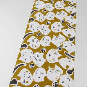 May include: A mustard yellow fabric with a repeating pattern of white cat faces with black eyes and noses. The cats have different expressions, including smiling, frowning, and surprised. The fabric also has small black paw prints scattered throughout.