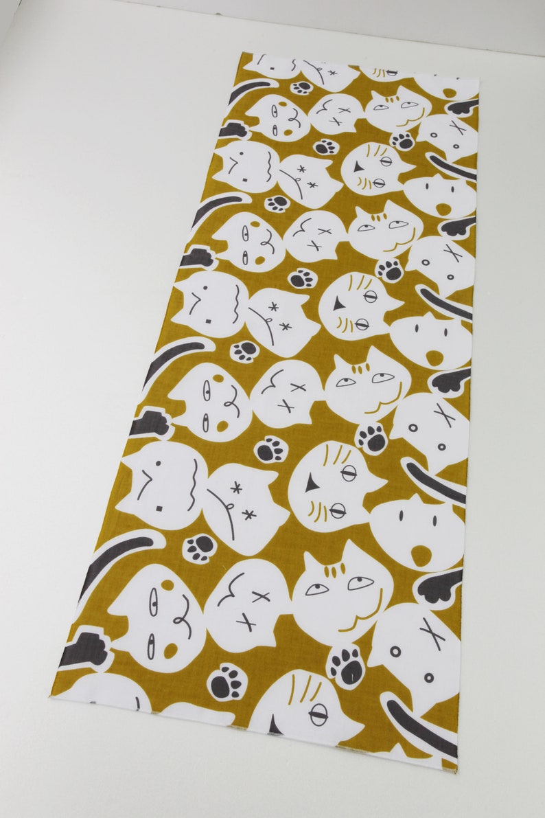 May include: A mustard yellow fabric with a repeating pattern of white cat faces with black eyes and noses. The cats have different expressions, including smiling, frowning, and surprised. The fabric also has small black paw prints scattered throughout.