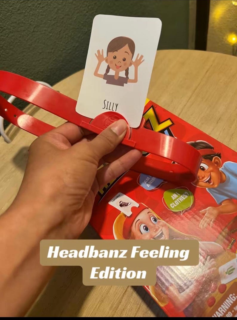 May include: A red plastic headband from the Headbanz Feeling Edition game, holding a white rectangular card with an illustration of a person making a silly face and the word SILLY printed below. The game box is visible underneath with colorful graphics.
