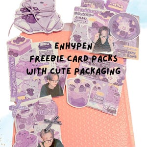 May include: A collection of purple-themed Enhypen freebie card packs with cute packaging, featuring illustrations of rabbits, berry desserts, and photographs of a person wearing glasses, all arranged on a light pink bubble mailer envelope.