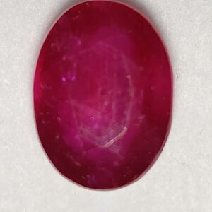 May include: A loose oval-shaped gemstone with a deep, saturated magenta-red color. The stone features a smooth, polished surface with visible internal facets and light reflections that highlight its translucent quality and vibrant red hue.