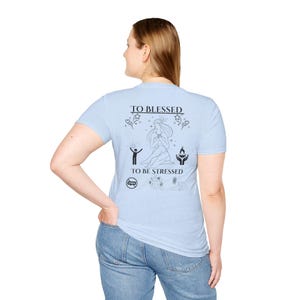 May include: A light blue t-shirt featuring a black graphic print on the back. The design includes the text TO BLESSED TO BE STRESSED, a central figure, stars, hearts, a stick figure with raised arms, a flame in hands, and small circular icons.