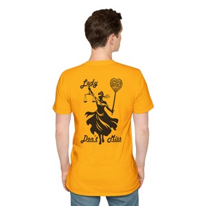 May include: A bright yellow short-sleeved t-shirt featuring a black graphic on the back. The design depicts a blindfolded figure holding scales and a heart-shaped sign with the word Justice, accompanied by the text Lady Don't Miss.