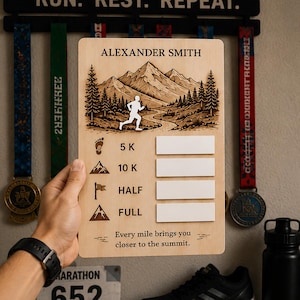May include: A wooden running medal display board featuring a mountain landscape and the name "ALEXANDER SMITH". It includes spaces for 5K, 10K, Half, and Full marathon medals, with the text "Every mile brings you closer to the summit."