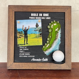 May include: A wooden framed display featuring a photo of a golfer, a 3D topographical map of a golf hole, and a white golf ball. Text reads: HOLE IN ONE, PEBBLE BEACH GOLF LINKS, HOLE #7, 106 YARDS, PAR 3, CLUB USED: PITCHING WEDGE, Alexander Smith.