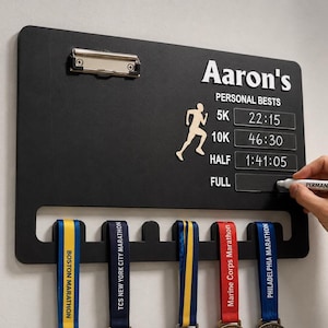 Pode incluir: Um expositor de medalhas retangular preto para parede com um clipe de metal no topo, uma silhueta branca de um corredor e texto branco Aaron's PERSONAL BESTS com espaços para tempos 5K, 10K, HALF e FULL, além de cinco ganchos para pendurar.