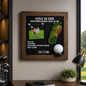 Pode incluir: Uma moldura de madeira quadrada contendo um painel preto com o texto HOLE IN ONE, SOUTHERS MARSH GOLF CLUB, HOLE #5, 138 YARDS, CLUB USED: PITCHING WEDGE, JULY 18, 2026. Inclui uma foto de três golfistas, um mapa de campo de golfe e uma bola.