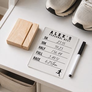 May include: A clear acrylic running personal best board with the text "ALEX'S PERSONAL BEST" and running times. A wooden block and a black and white marker are next to the board. Running shoes are in the background.