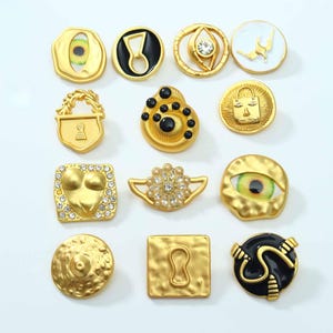 May include: A collection of twelve decorative metal buttons in various shapes and designs, including gold-toned finishes, black enamel accents, clear rhinestones, and eye-themed motifs. The buttons feature diverse textures, geometric forms, and symbols.
