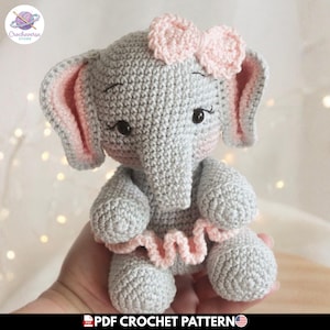 Puede incluir: Un juguete de elefante tejido a ganchillo en color gris con un lazo rosa claro en la cabeza, orejas interiores rosas y una falda con volantes rosa en la cintura. El elefante tiene ojos negros bordados con pestañas largas y trompa. Texto: PDF CROCHET PATTERN.