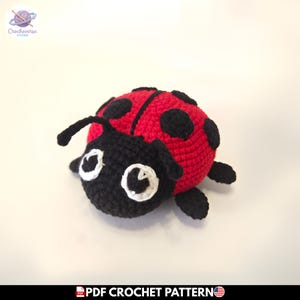 Puede incluir: Un juguete de mariquita tejido a ganchillo a mano con un cuerpo redondeado de color rojo brillante y puntos negros. La cabeza es negra con dos antenas curvas y ojos grandes delineados en hilo blanco. Tiene pequeñas patas negras a los lados.