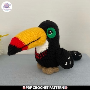 Puede incluir: Un pájaro tucán de ganchillo hecho a mano con un gran pico amarillo con puntas rojas y negras, cuerpo negro, pecho blanco, parches verdes en los ojos y patas de color marrón claro. El texto PDF CROCHET PATTERN aparece en la parte inferior.