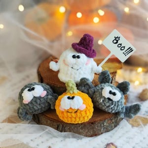 Puede incluir: Un conjunto de cuatro figuras de Halloween tejidas a ganchillo que incluye un fantasma blanco con sombrero morado y un cartel que dice Boo!!!, una araña gris, una calabaza naranja y un murciélago gris, todos con ojos blancos y mejillas rosas.