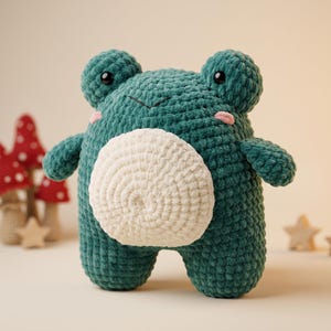 Puede incluir: Un peluche de rana tejido a ganchillo en color verde oscuro con una gran barriga redonda de color crema. El juguete cuenta con dos ojos prominentes, pequeñas pupilas negras y dos parches bordados en rosa claro en las mejillas, hecho de hilo suave.