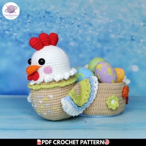 Puede incluir: Una cesta de ganchillo con forma de gallina, cabeza blanca, cresta y barbillas rojas, pico naranja y ojos negros. El cuerpo es color canela con detalles pastel, un ala con volantes verdes y azules, y una cesta llena de huevos moteados.