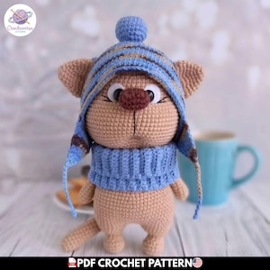 May include: A crocheted cat doll wearing a blue and brown striped hat and scarf. The cat has a brown nose and large white eyes. The doll is tan and has a long tail. The image includes the text "PDF Crochet Pattern".