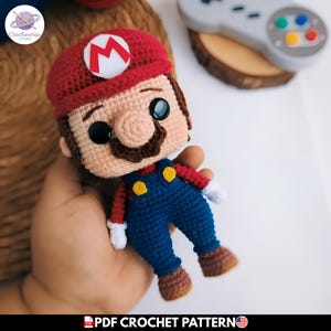 Puede incluir: Muñeco de Mario tejido a ganchillo, con gorro rojo con círculo blanco y "M" rojo, peto azul y bigote marrón. El muñeco tiene ojos negros y sostiene un mando. El texto "PDF CROCHET PATTERN" está en la parte inferior.