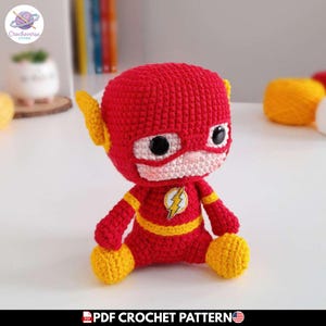 Puede incluir: Una muñeca de ganchillo roja y amarilla con una cabeza grande, ojos de plástico negro y un emblema de rayo en el pecho. La figura tiene alas amarillas en las orejas y detalles amarillos en brazos, cintura y pies. El texto inferior dice PDF CROCHET PATTERN.