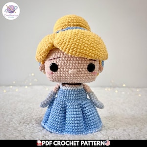 Puede incluir: Una muñeca de ganchillo que representa un personaje con cabello amarillo brillante recogido en un moño, vistiendo un vestido azul claro con guantes largos a juego. La muñeca tiene tez beige, grandes ojos de botón negros y pendientes de perlas.