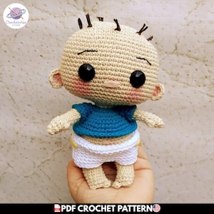 Puede incluir: Una muñeca de ganchillo con cabeza beige, ojos de botón negros y cabello castaño oscuro escaso. La muñeca viste una camiseta azul de manga corta y pantalones cortos blancos con detalles amarillos. Tiene mejillas rosadas y boca bordada.