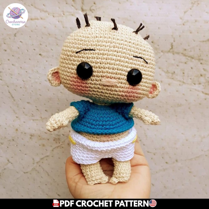 May include: A crocheted doll with a beige head, black button eyes, and sparse dark brown hair. The doll wears a blue short-sleeved shirt and white shorts with yellow accents. The doll has pink blush on its cheeks and a simple stitched mouth.