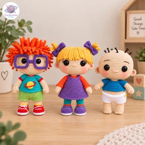 May include: Three handmade crochet dolls featuring characters with distinct features. The left doll has orange hair, purple glasses, and a teal shirt with a planet graphic. The center doll has yellow hair with purple bows and a purple dress. The right doll is bald with a blue shirt and white shorts.