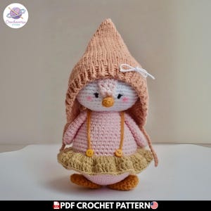 Puede incluir: Una muñeca pingüino de ganchillo que lleva una capucha puntiaguda de color melocotón con un lazo blanco, un cuerpo rosa claro y una falda beige con volantes y tirantes amarillos. El pingüino tiene cara blanca, ojos negros, pico amarillo y mejillas rosadas.