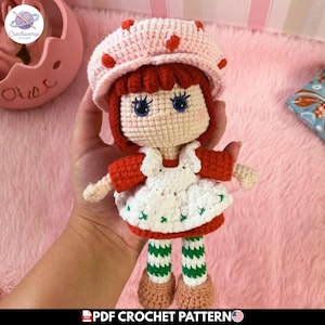 Puede incluir: Una muñeca de ganchillo hecha a mano con un sombrero rosa claro con detalles de fresas rojas, pelo rojo oscuro y ojos azules. La muñeca lleva un delantal blanco sobre un vestido rojo, piernas a rayas verdes y blancas y zapatos marrones. Texto: PDF CROCHET PATTERN.