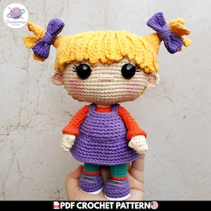 Puede incluir: Una muñeca tejida a mano con cabello amarillo brillante peinado en dos coletas con lazos morados, ojos negros grandes y mejillas rosadas. La muñeca viste una camisa naranja de manga larga bajo un vestido morado, con leggings verde azulado y zapatos morados.