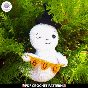 Puede incluir: Un fantasma amigurumi de ganchillo blanco con un sombrero puntiagudo negro. El fantasma tiene un ojo guiñando y un ojo de cuenta negra, y sostiene una pequeña pancarta naranja con el texto negro BOO impreso en las banderas triangulares.