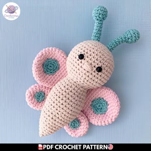 Puede incluir: Un juguete de mariposa hecho a ganchillo a mano con cabeza y cuerpo de color rosa pálido, dos antenas de color verde azulado con puntas redondeadas y cuatro alas rosas decoradas con acentos circulares verde azulado. Tiene dos pequeños ojos negros.