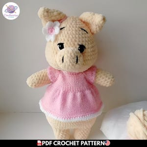 Puede incluir: Un peluche de cerdo tejido a ganchillo en color crema con un acabado de hilo suave y texturizado. El cerdo lleva un vestido tejido de color rosa claro con un borde blanco en el dobladillo y una pequeña flor blanca con centro rosa en la oreja.