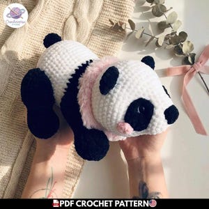 Puede incluir: Un peluche de panda tejido a ganchillo con cuerpo blanco y negro, orejas negras, parches negros alrededor de los ojos y una pequeña nariz negra. El panda tiene un cuello de piel sintética rosa claro y un pequeño detalle de rubor rosa en la mejilla.