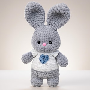 Puede incluir: Un conejo de peluche tejido a ganchillo en color gris con orejas largas, ojos de plástico negro y una pequeña nariz rosa. El conejo lleva una camiseta blanca tejida con un corazón azul tejido en el centro del pecho, hecho de lana suave.