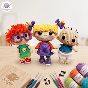 May include: Three crocheted dolls featuring characters with distinct hair and clothing. One has orange hair and purple glasses, one has yellow pigtails with purple bows, and one is bald. Includes crochet hooks, safety eyes, and yarn balls.