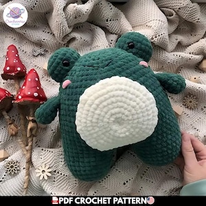 Puede incluir: Un juguete de peluche de rana verde oscuro con una barriga grande, redonda y texturizada en blanco, dos ojos negros pequeños y mejillas bordadas en rosa. La rana tiene una construcción suave de hilo con un patrón de ganchillo visible.