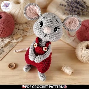 Puede incluir: Un juguete de ratón tejido a ganchillo con hilo gris claro, orejas grandes y redondas con centros rosados y una pequeña nariz rosa. El ratón viste un peto de color rojo oscuro con dos pequeños botones de madera en los tirantes y ojos negros.