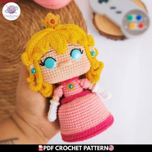 Puede incluir: Una muñeca de ganchillo hecha a mano de una princesa, con una corona y pelo amarillos, un vestido rosa y ojos azules. La muñeca se sostiene en una mano. Es un patrón de ganchillo en PDF.